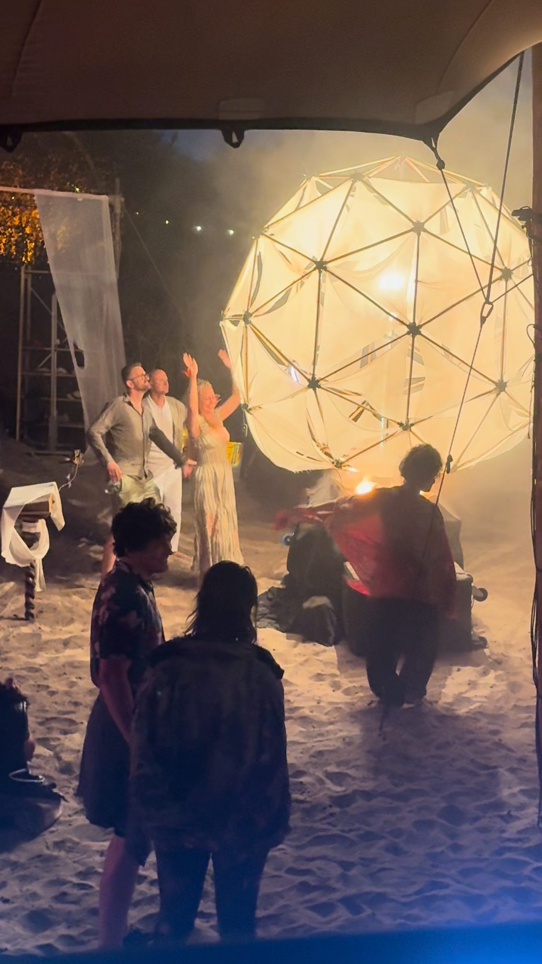 Sól fully awake at night — the bamboo orb glowing warm white through haze, people beside it on the sand.