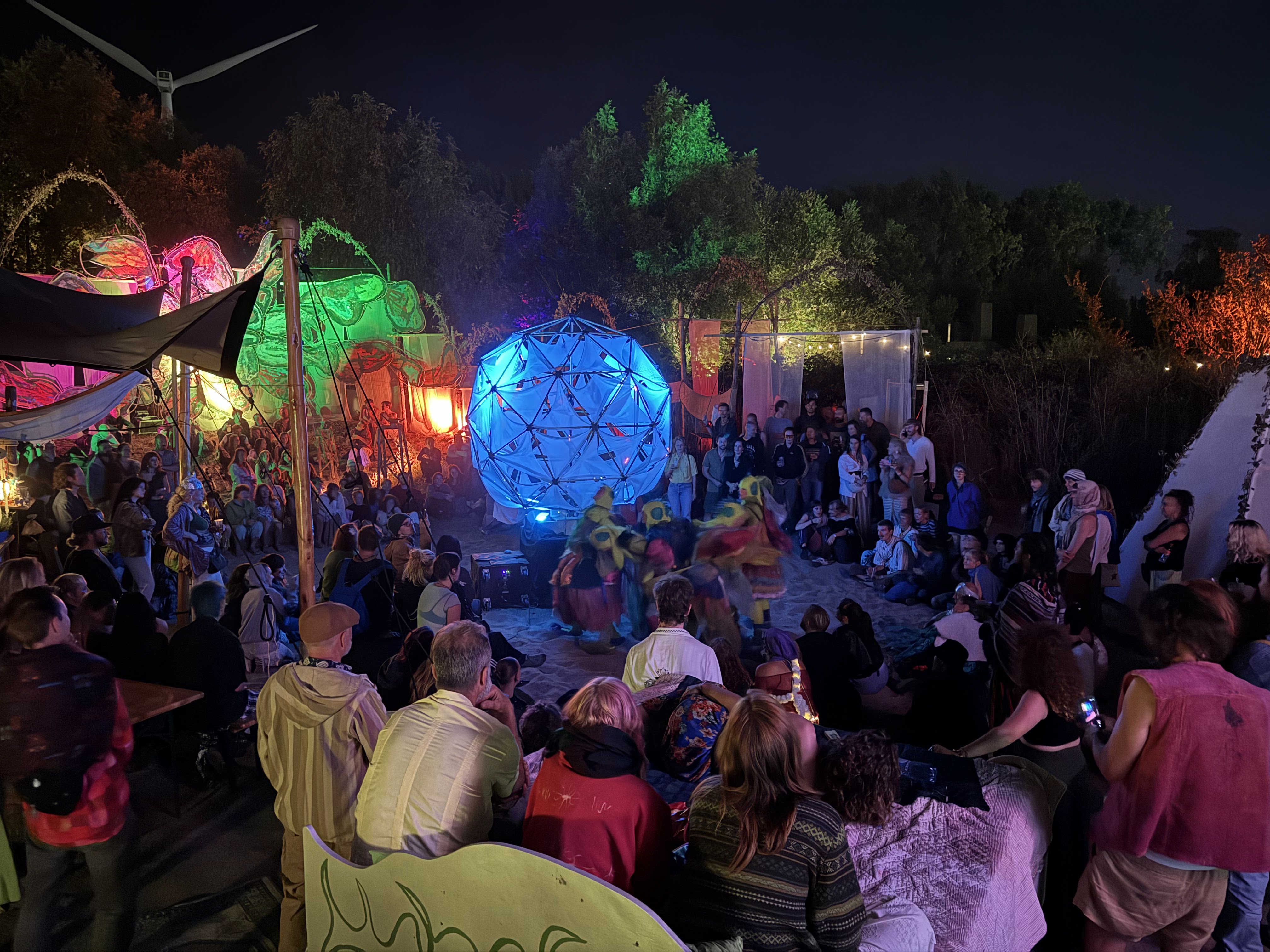 Sól awake at night at Het Landjuweel — the bamboo geodesic orb glowing blue on the sand, ringed by a seated crowd with dancers moving in front of it.